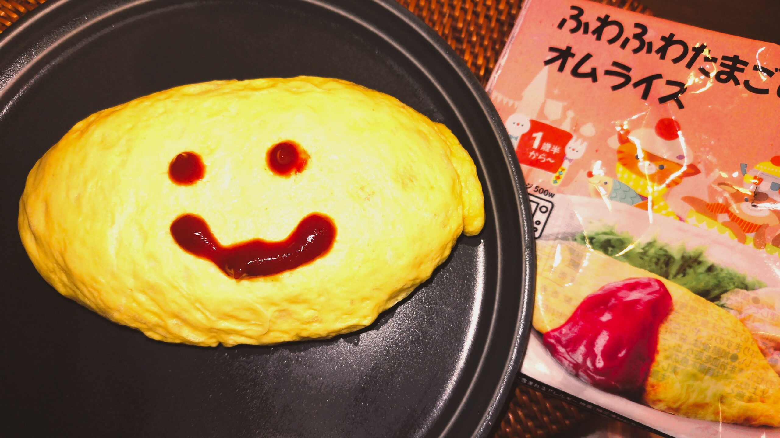 モグモのふわふわたまごのオムライスをお皿に出し、ケチャップで笑顔を描いたところ。右側に商品のパッケージ