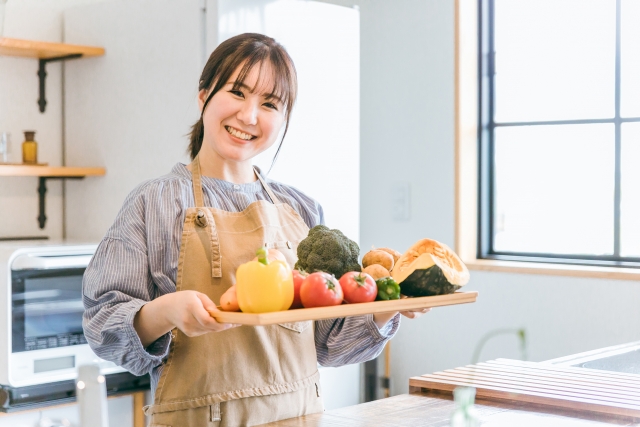 野菜をのせた木のトレーを持って笑うエプロン姿の女性。幼児食を監修する管理栄養士のイメージ